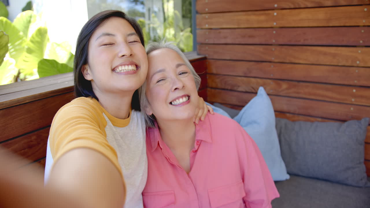 Taking selfie, young asian woman and senior asian woman smiling and hugging on couch