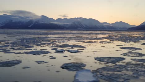 Stunning-aerial-over-icy-water-with-majestic-mountains-in-background-at-sunrise