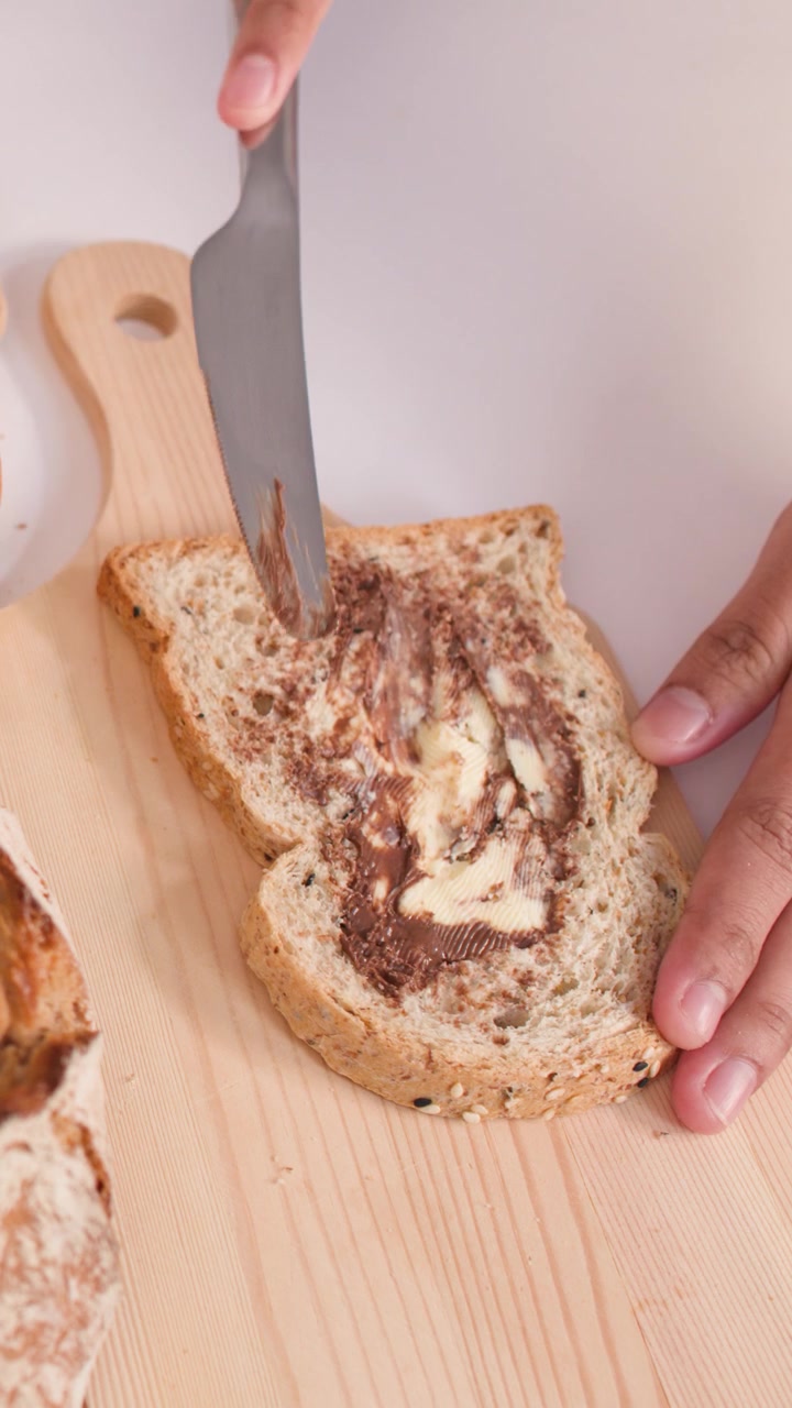 Overhead view of hands spreading creamy nut butter onto multigrain bread using a metal knife.