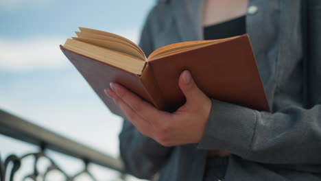 closeup of lady holding an open book with her left hand supporting it from beneath, standing close to an iron fence, the pages slightly lift as if from a gentle breeze