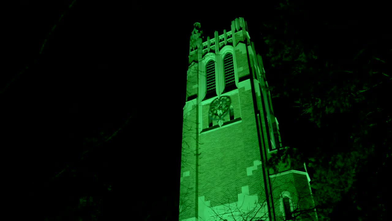 2023년 2월 대량 총격 사건의 희생자를 기리기 위해 밤에 미시간 주립 대학 캠퍼스에 있는 보몬트 타워가 나무 사이를 걷는 클로즈업 비디오와 함께 녹색으로 점등되었습니다.