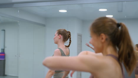 women engage in a workout session, removing hands from heads while performing a spine stretch, large mirror reflects the gym environment