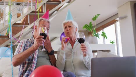 senior caucasian couple blowing party blowers celebrating birthday toasting wine glasses at home