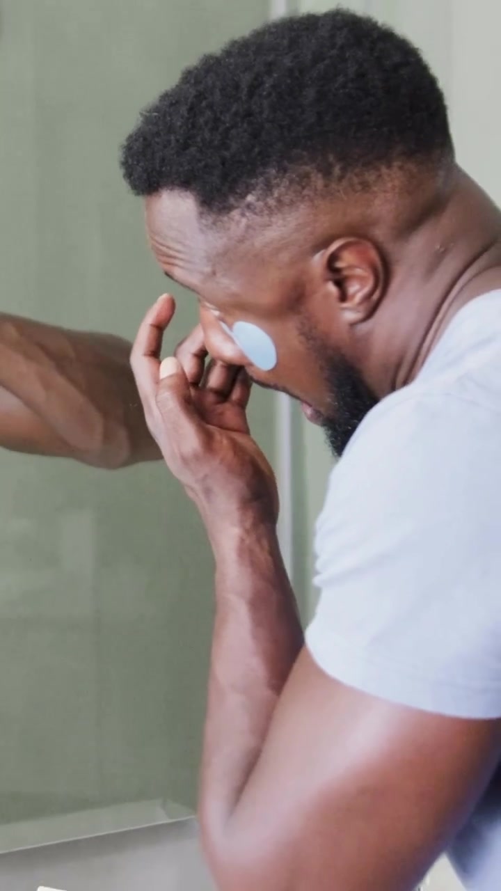 Vertical video: Starting skincare, man in gray tee pressing eye patch at home vanity, pink phone