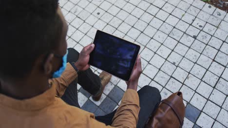 African-american-businessman-wearing-face-mask-using-tablet-in-sitting-in-street