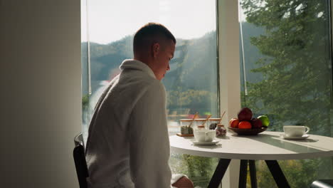 young man moves chair to table in morning. husband comes to kitchen to have healthy breakfast by window with view on nature. morning rituals during rest time