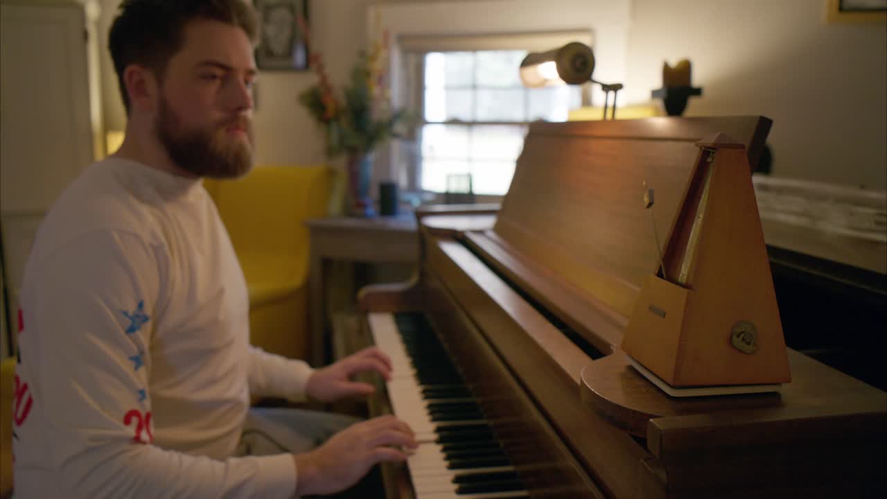 un joven barbudo practica la música del piano en su sala de estar usando un metrónomo de madera antiguo rústico que balancea su péndulo a 60 bpm