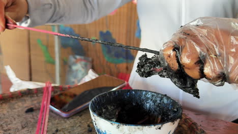 a gloved hand dips an incense stick into a thick black herbal paste as part of the traditional incense-making process in quang phu cau village, ha noi, vietnam, with tools and raw materials in view.