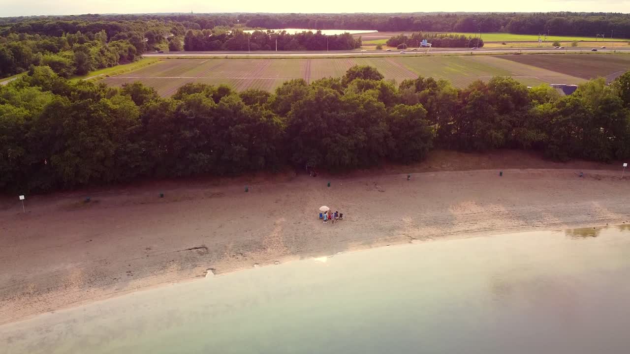 Aerial view above a lake beach with multicultural friends hanging out, distant agrilcural fields and highway near Breda, Netherlands.