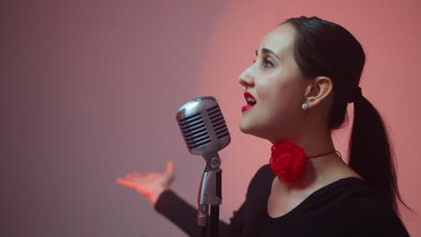 primer plano de una cantante apasionada con labios rojos y una cinta roja en el cuello, cantando en un micrófono vintage, mientras mueve sus manos expresivamente, la cámara se aleja lentamente