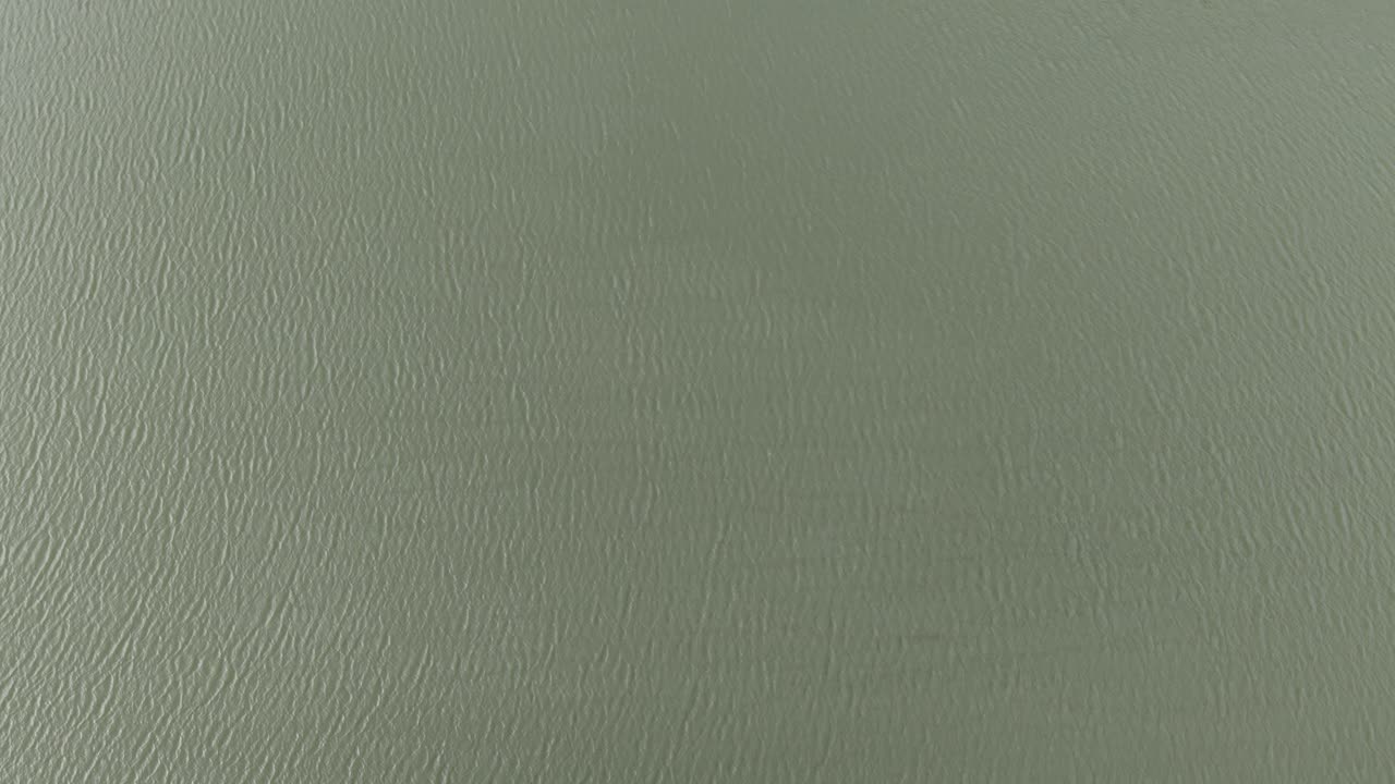 Aerial view of a lake shore lined with vibrant autumn trees. The brilliant red, orange, and green foliage contrasts beautifully with the calm, rippled surface of the water