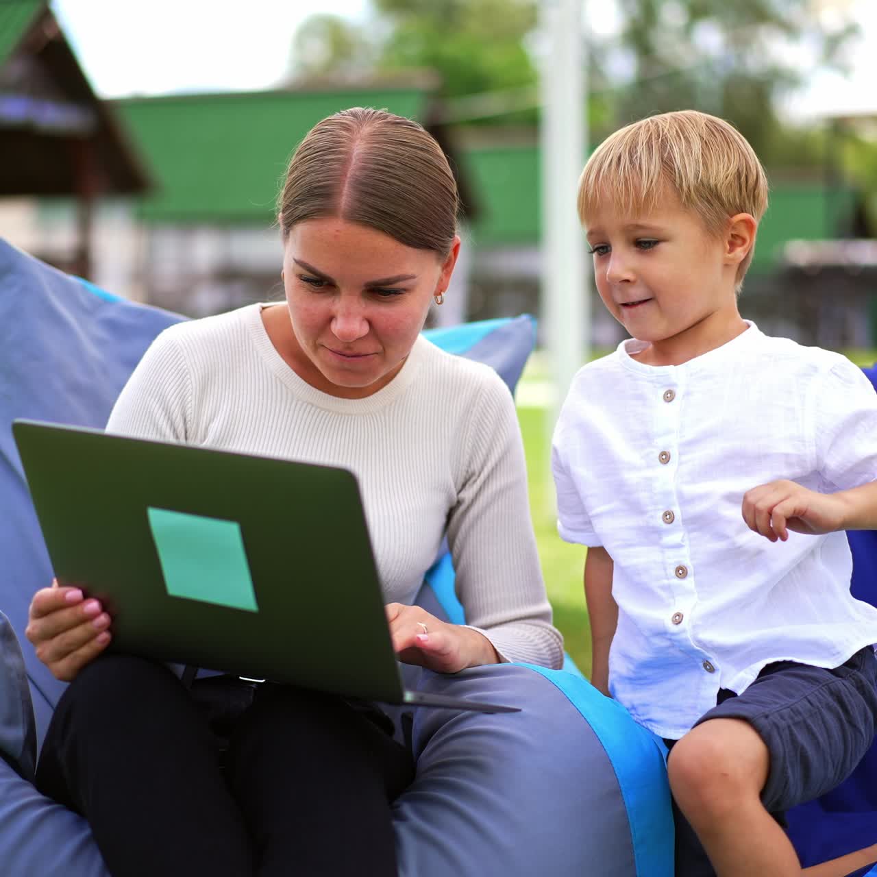 Woman and boy sitting in bean bag chairs looking at laptop, laughing and playing cheerfully. Mom and son discussing cartoons. Blurred backdrop