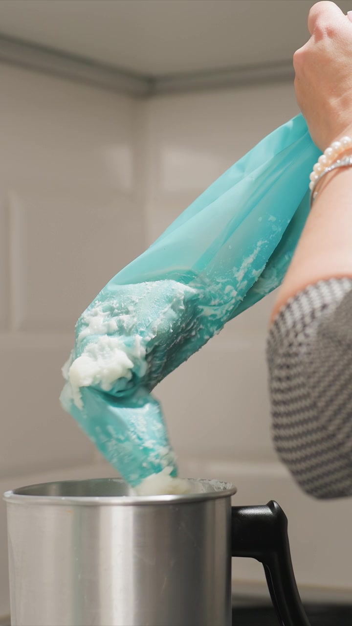 Primo piano di una persona che indossa maniche grigie testurizzate e braccialetti di perle, mentre versa sapone blu in un contenitore di metallo argentato. Il sapone è turchese con spirali bianche; lo sfondo presenta una cucina neutra.