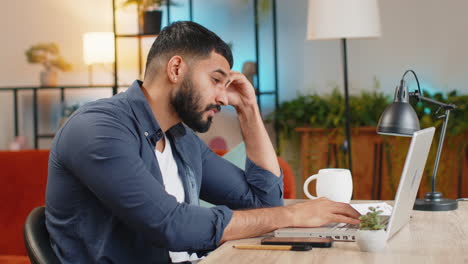 Bored-sleepy-young-Indian-man-working-on-laptop-computer-yawning-leaning-on-hand-at-home-office