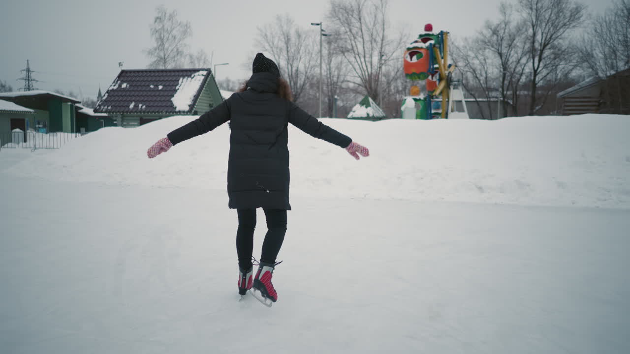 Skater in black coat and red skates gliding on outdoor ice rink during cold winter season, arms outstretched for balance, snow surrounding area with bare trees and distant colorful amusement structure