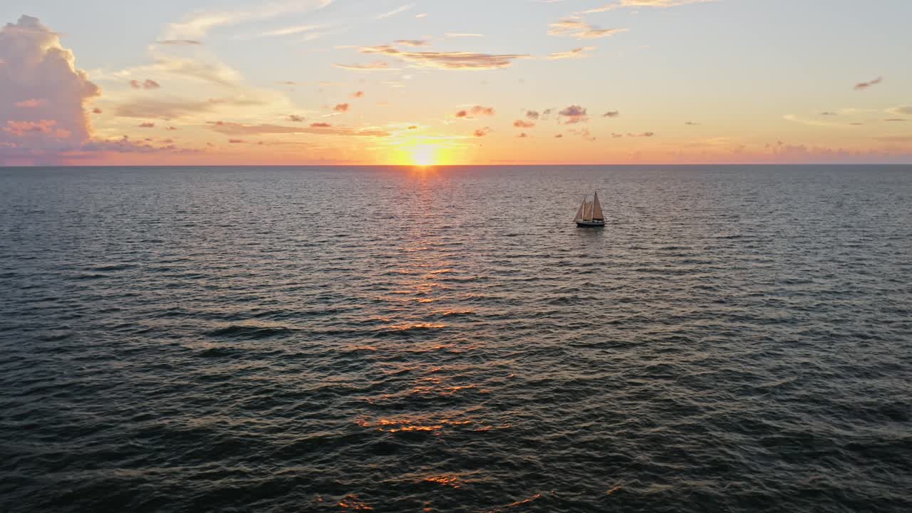 A sailboat glides toward the horizon as the sun sets, casting soft golden and orange hues across the sky and shimmering reflections over rippling ocean waters