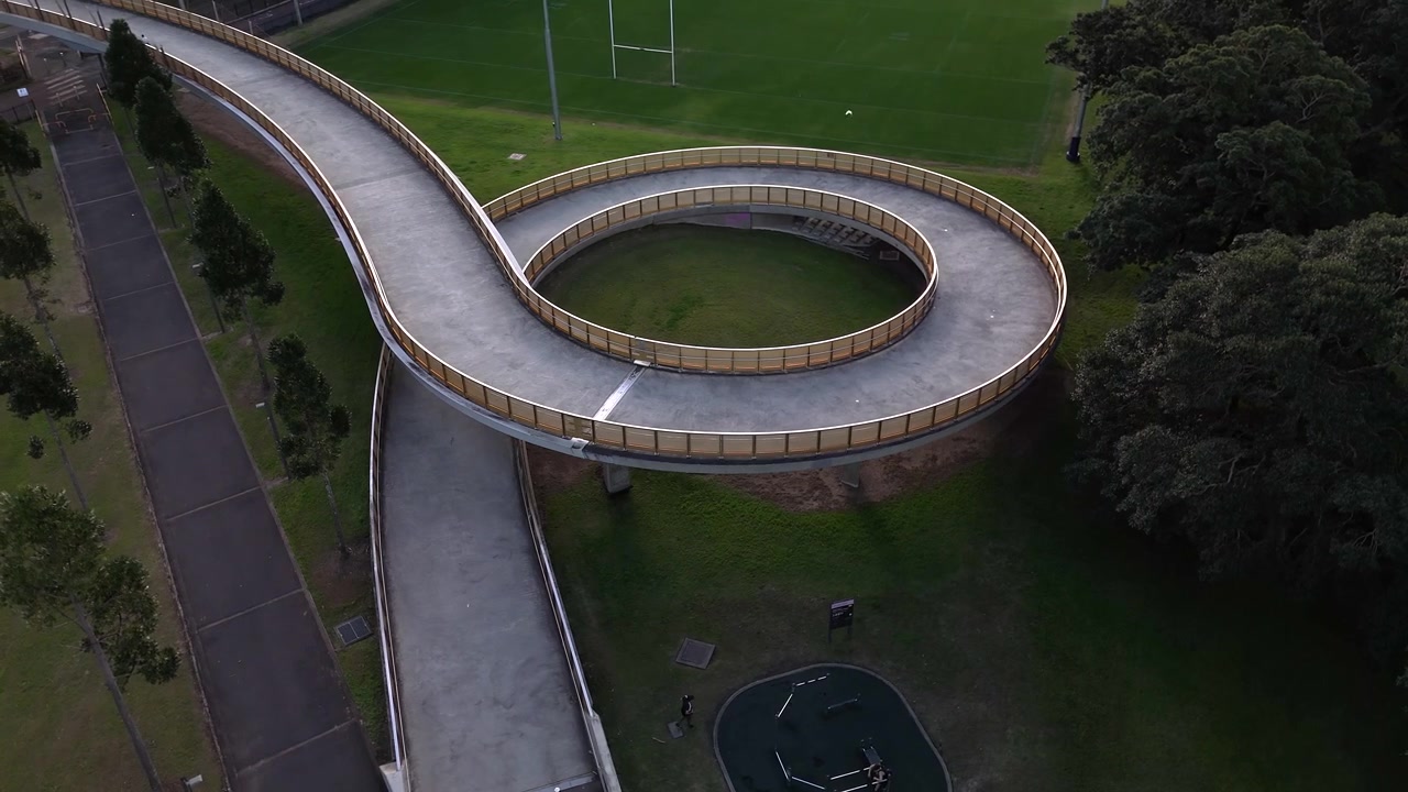 Vista aérea de uma grande ponte em espiral transportando pedestres, ciclistas e corredores. A câmera orbita a estrutura.