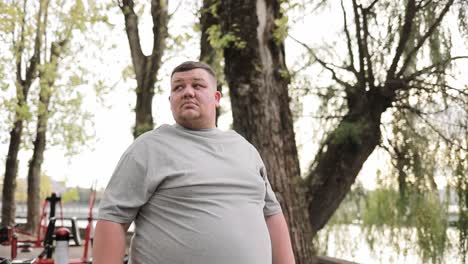portrait of an overweight young man standing against the background of trees in the park portrait of an overweight young man standing against the background of trees in the park a fat man in a gray tshirt in a green zone near outdoor exercise machines