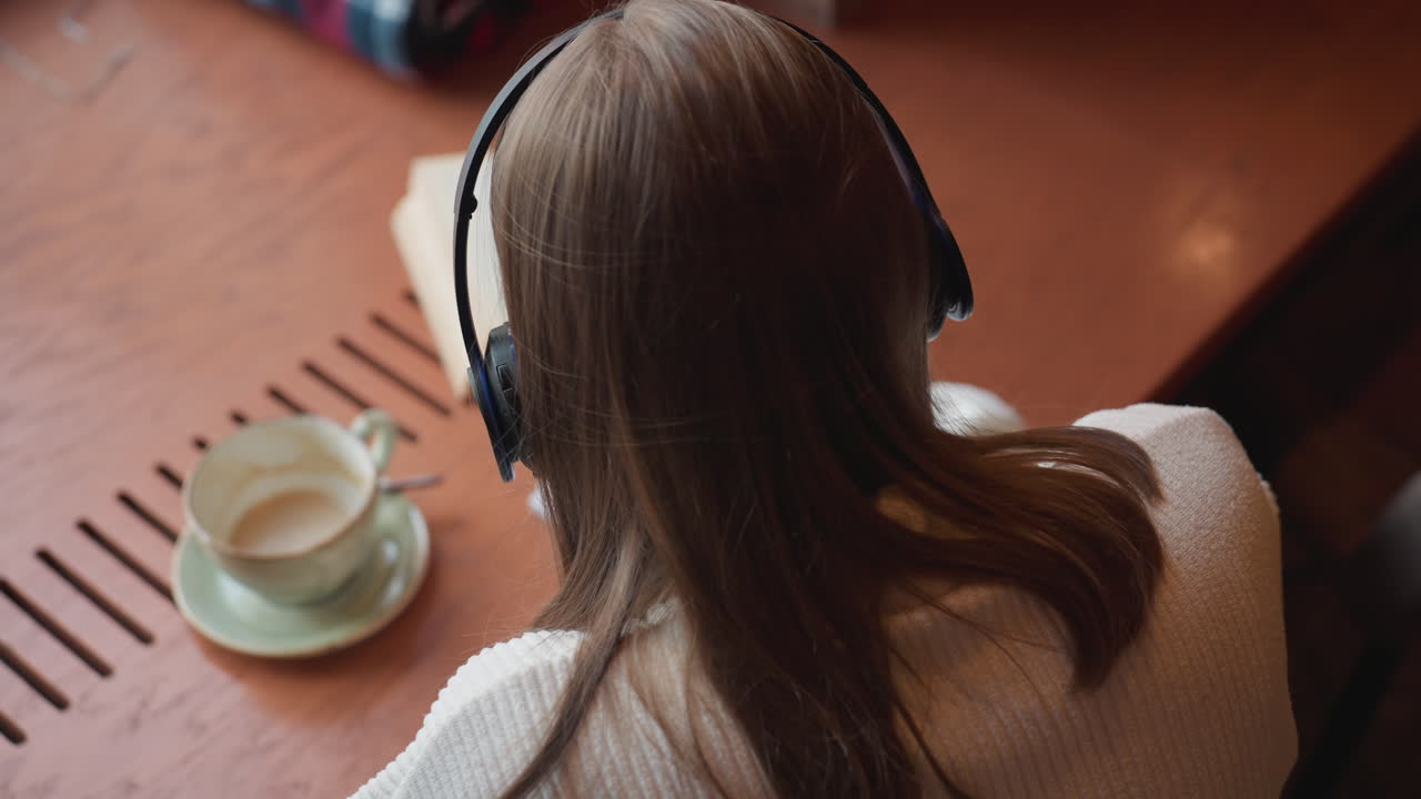 back view of female lecturer with headphones writing in notebook while listening to music beside open book and coffee cup on wooden desk in cozy room with warm lighting and focused atmosphere
