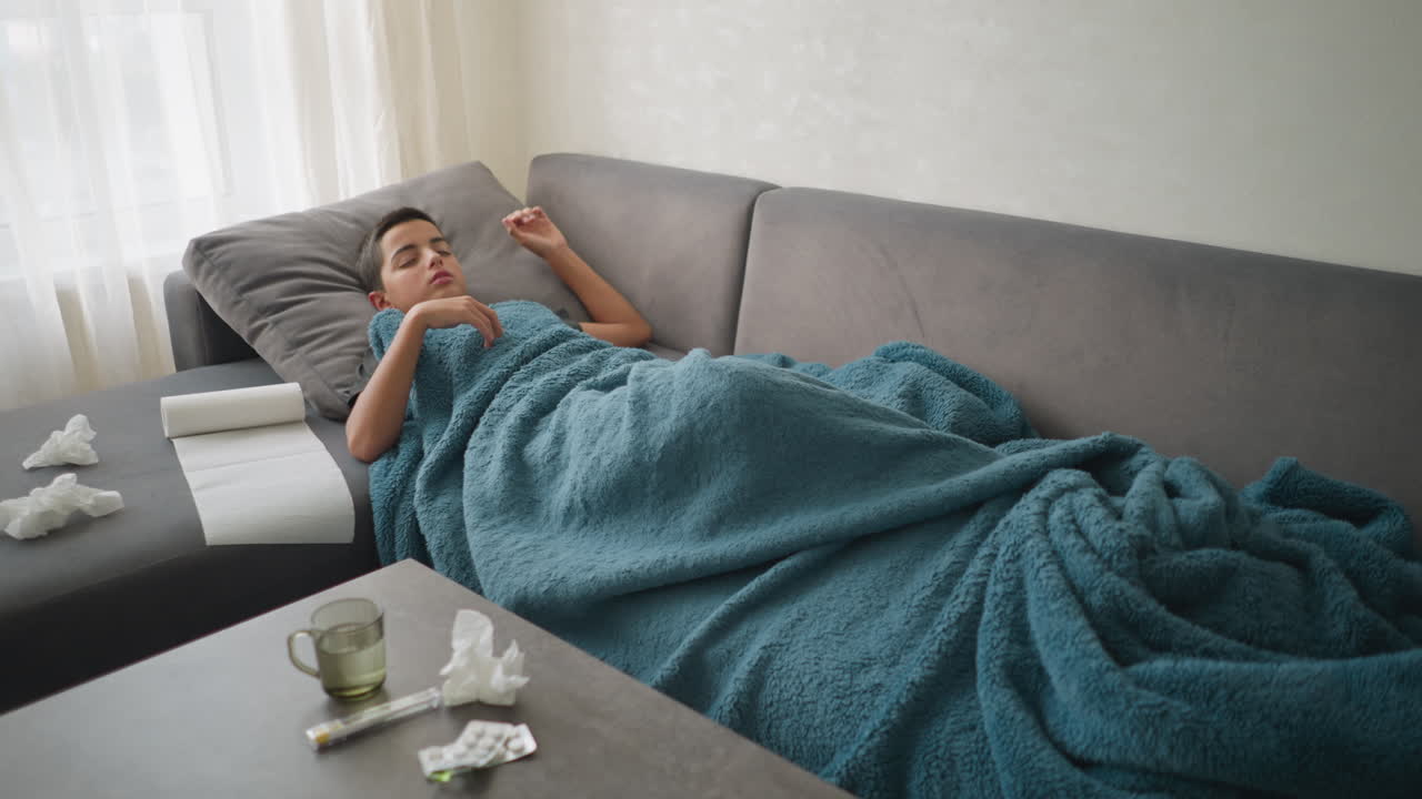 Scattered room with boy lying on couch under blanket, adjusting while glass cup and tissue papers are on the table, messy environment showcasing a sick child recovering at home