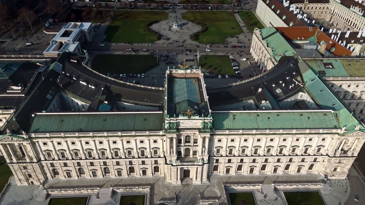 Panoramatický letecký záběr dronem nad palácem Hofburg zezadu, směřující na zámecký areál ve Vídni, zobrazující okolní panorama města, zimní sluneční světlo, zataženou oblohu a turisty procházející se po areálu