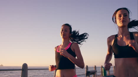 Zwei-Sportliche-Frauen-Laufen-Im-Freien-In-Zeitlupe-Auf-Der-Promenade-Bei-Sonnenuntergang-Am-Meer-Und-Genießen-Den-Abendlauf