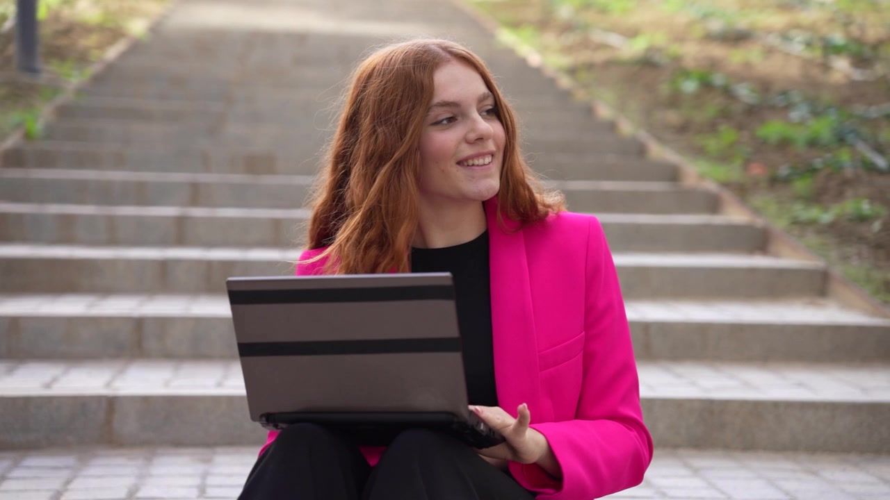 Una giovane donna seduta all'aperto che usa il suo laptop, vestita con un blazer rosa acceso.
