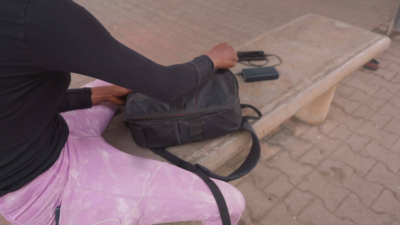 woman sitting on bench with backpack, reaching for phone on stone bench pink skirt and black top, hands adjusting zipper, urban paved ground, candid commuter moment, daylight waiting scene focused