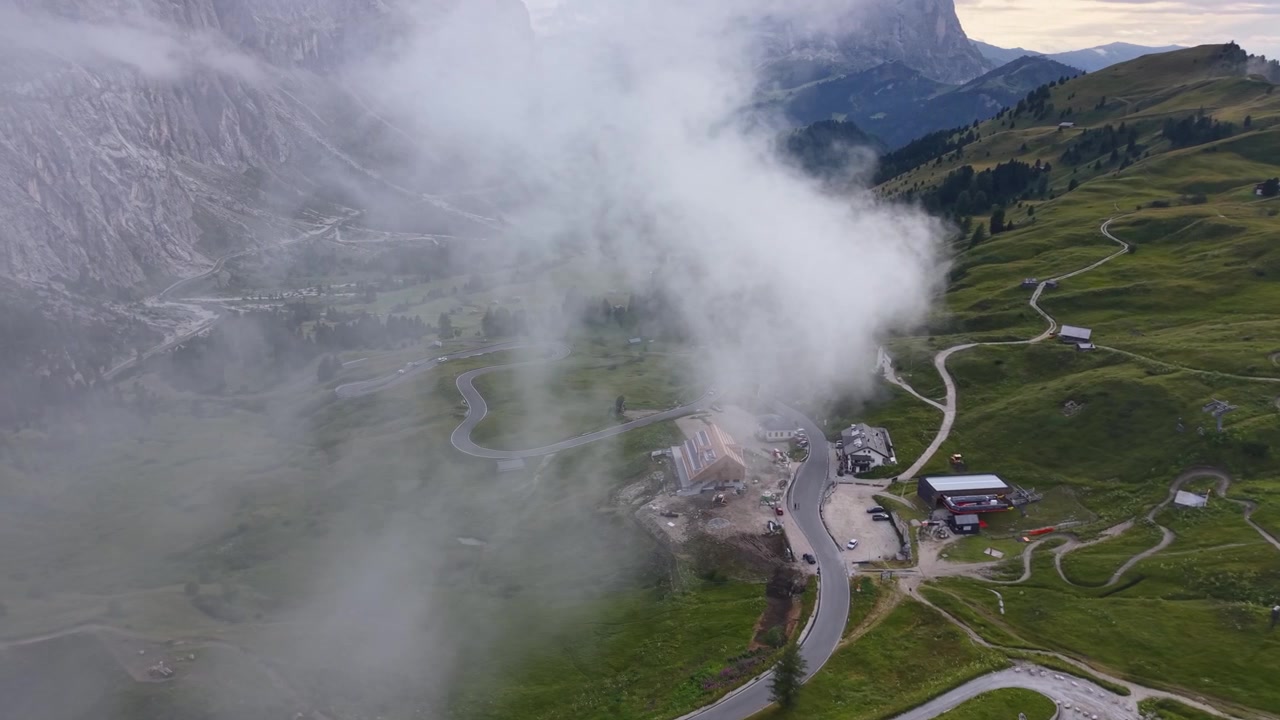 Veduta cinematografica con drone sopra il Passo Gardena, mentre nuvole alla deriva nascondono parzialmente strade alpine tortuose e aspri paesaggi montani nelle Dolomiti, Italia.