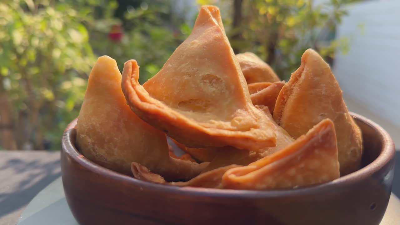 Primo piano rotante di un piatto di croccanti samosa dorate impilate in una ciotola, bordi di pasta sfoglia che catturano la calda luce del sole mentre un movimento delicato rivela la consistenza e la forma