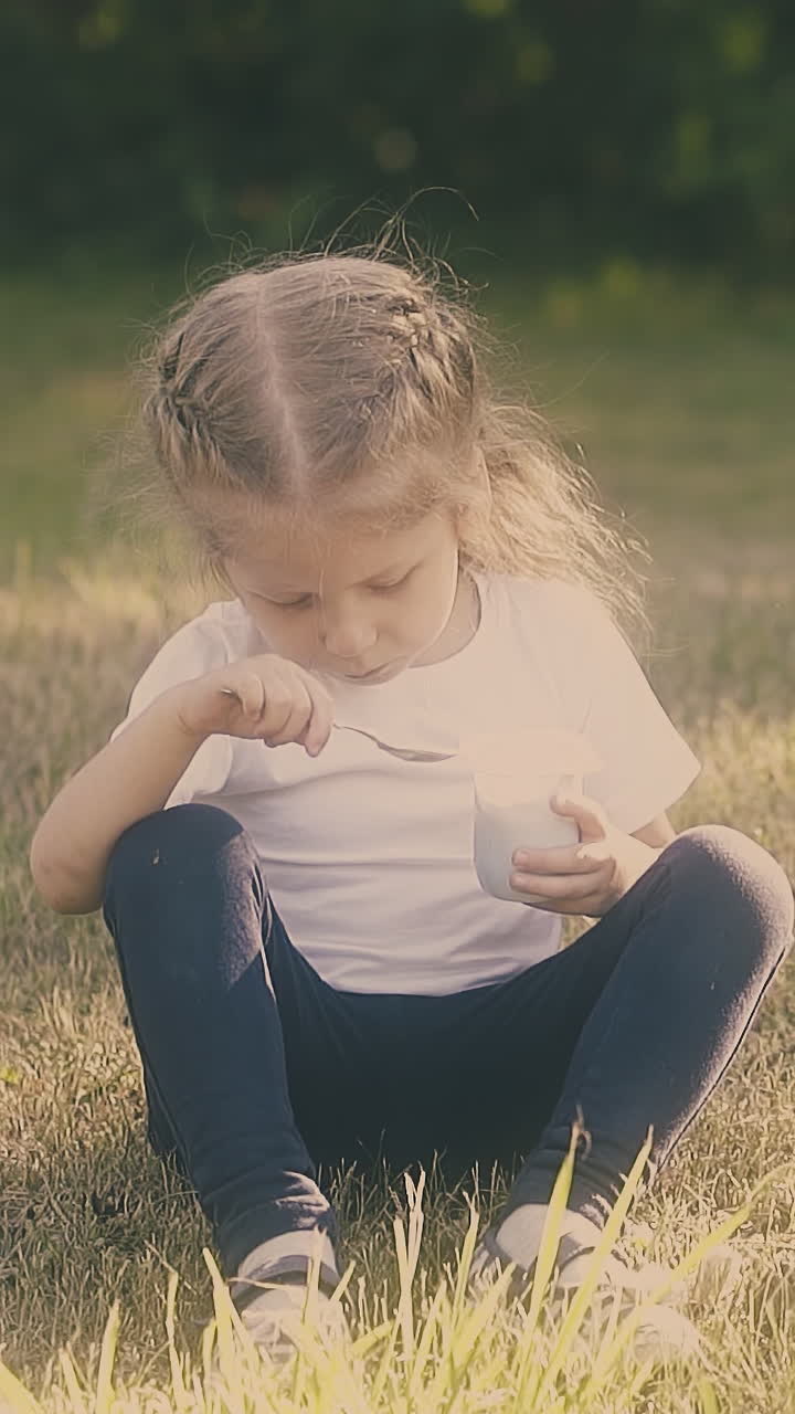 girl with braids wearing white t-shirt eats tasty dessert holding white cup and sitting on yard lawn closeup slow motion