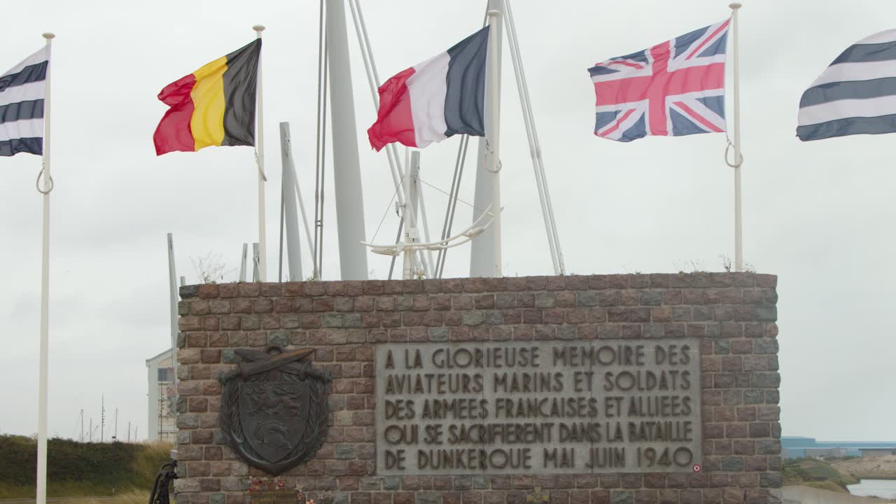 Stone war memorial with waving Allied flags, overcast daylight, static camera, coastal environment