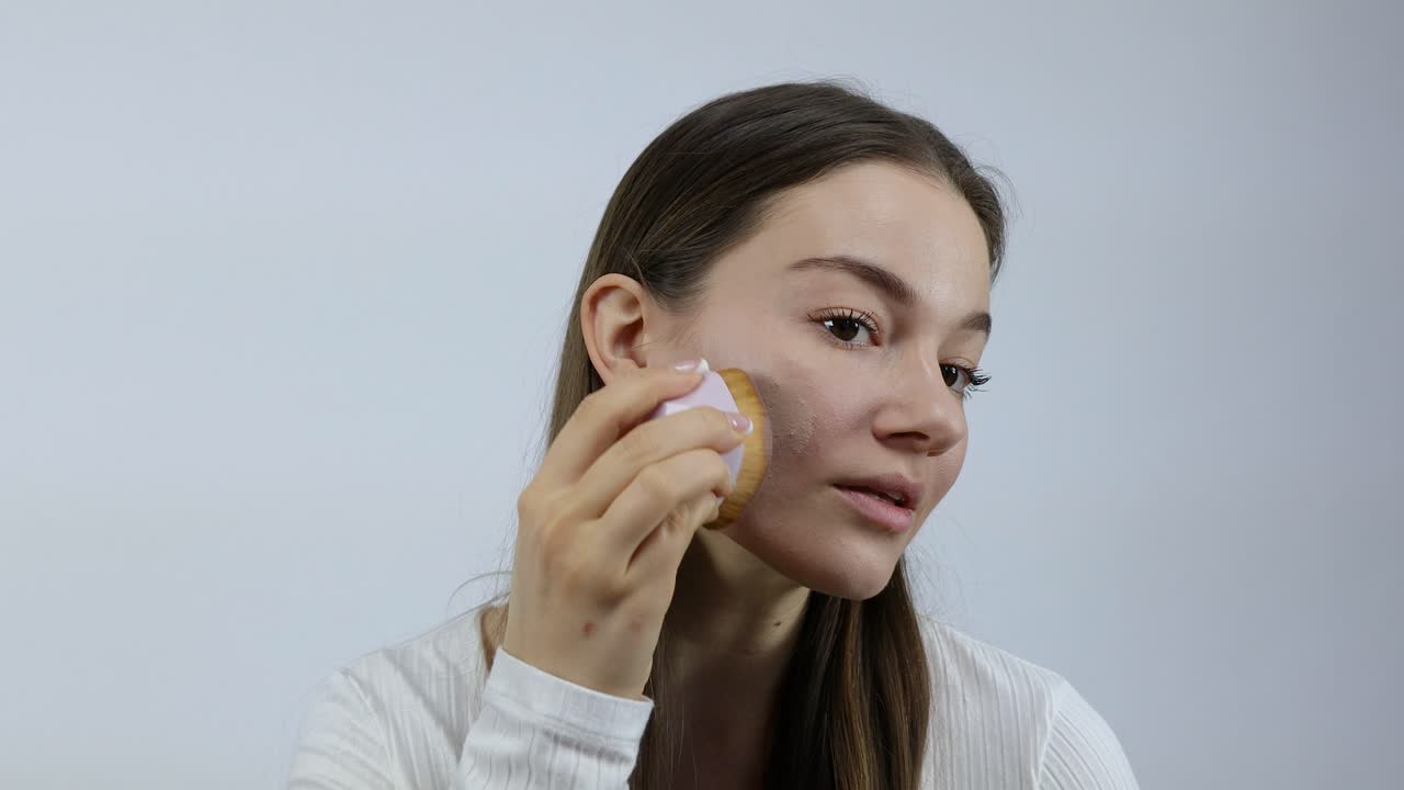 아름다운 젊은 유럽 여성이 색 배경 앞에 을 사용하여 얼굴에 메이크업을 적용하고 퍼뜨리고 있습니다.