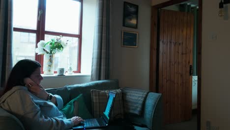 Woman-relaxing-on-a-sofa-using-her-mobile-smartphone-and-working-on-her-laptop