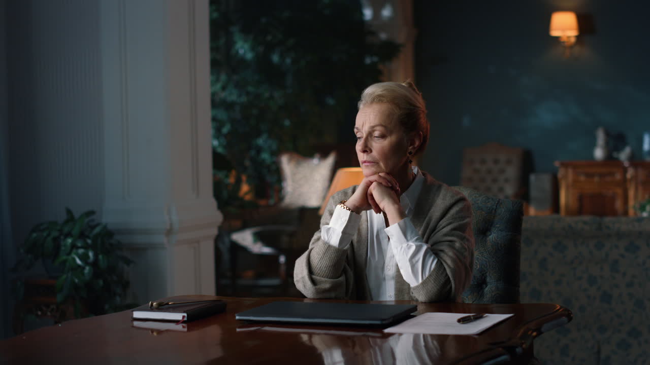 Thoughtful senior woman sitting at table in luxury cabinet