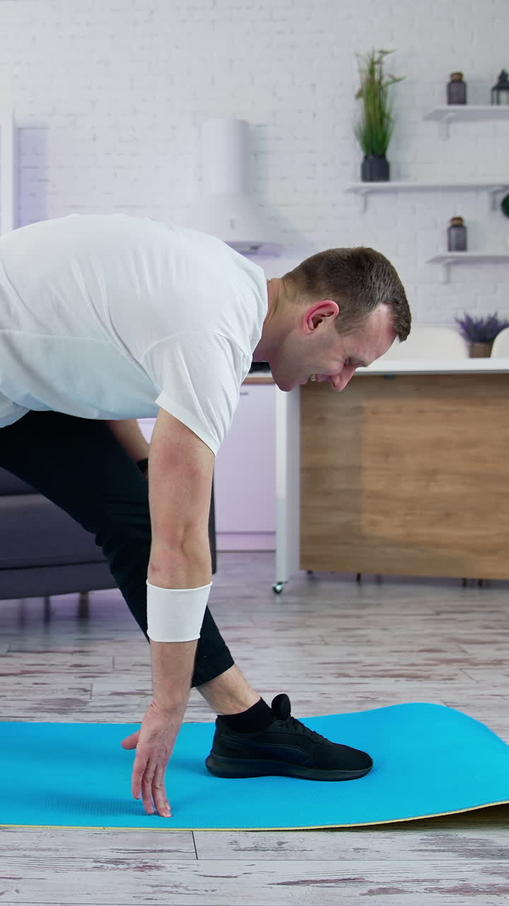 Young man doing fitness at home. Man in t-shirt and pants is doing exercises on mat at home in his spacious kitchen Vertical video