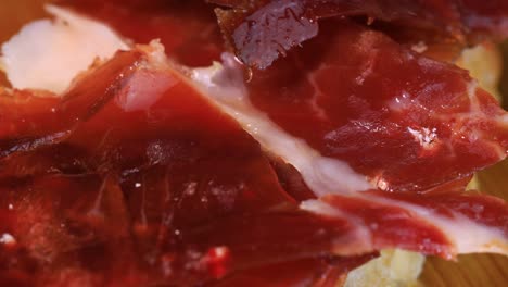 macro shot of oily high quality spanish serrano ham with bread rotating on wooden board, close up view from above