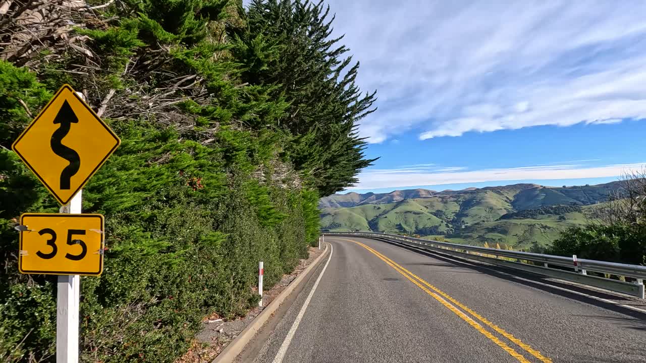 An ambulance travels through a winding road in Akaroa, New Zealand, under clear skies and lush greenery