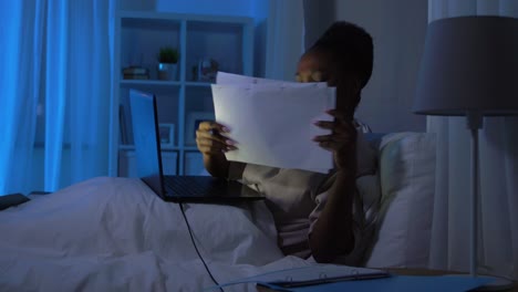 African-Woman-with-Laptop-Working-in-Bed-at-Night.technology,-remote-job-and-people-concept-happy-smiling-young-african-american-woman-with-wireless-earphones,-laptop-computer-and-papers-working-in-bed-at-home-at-night