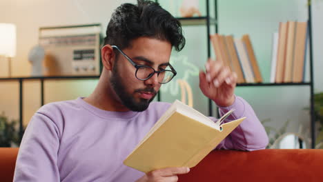 Hombre-Indio-Leyendo-Un-Libro-Interesante-Pasando-Páginas-Sonriendo-Disfrutando-De-La-Literatura-Tomando-Un-Descanso-En-El-Sofá