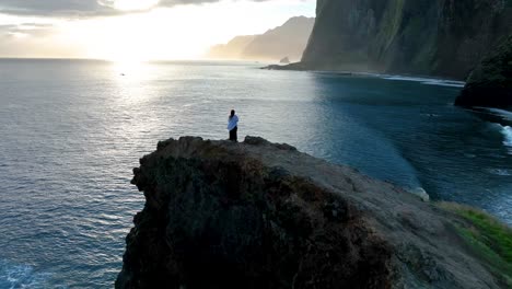 Aerial-flyover-of-woman-standing-on-coastline-rock-cliff-with-drone-in-the-sky,-Madiera