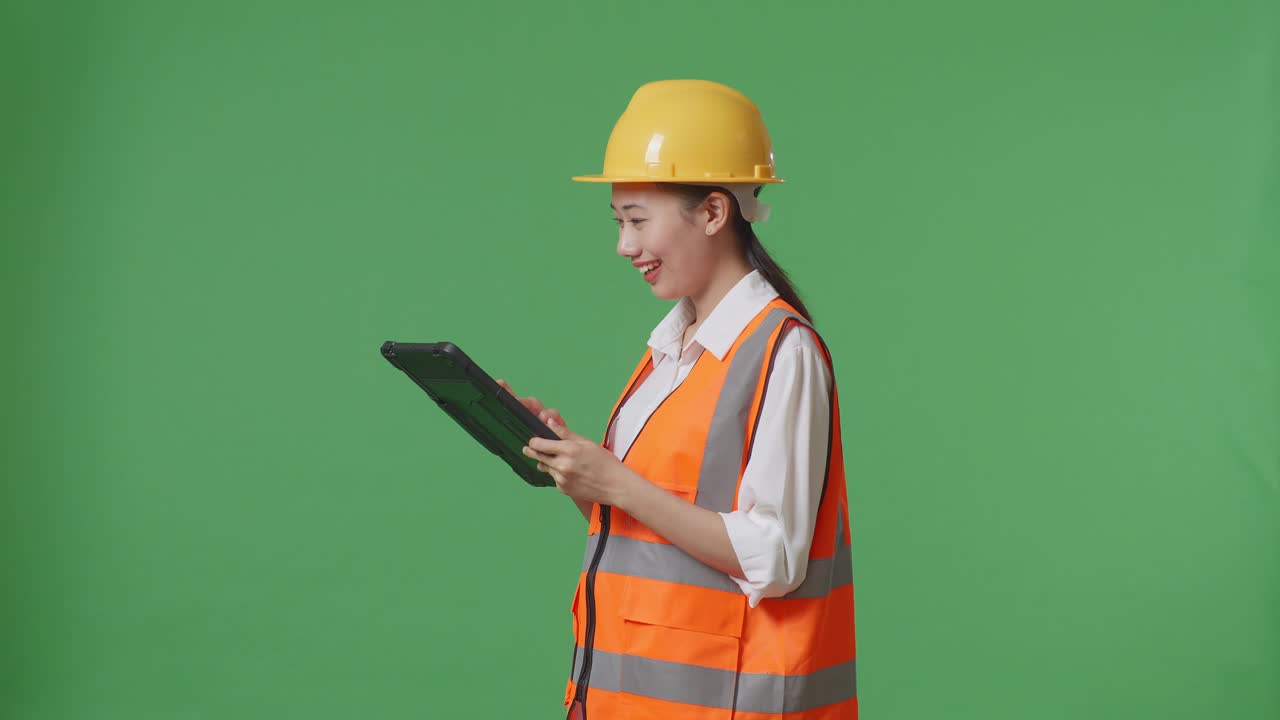 Side View Of Asian Female Engineer With Safety Helmet Looking At The Tablet In Her Hand And Looking Around While Standing In The Green Screen Background Studio