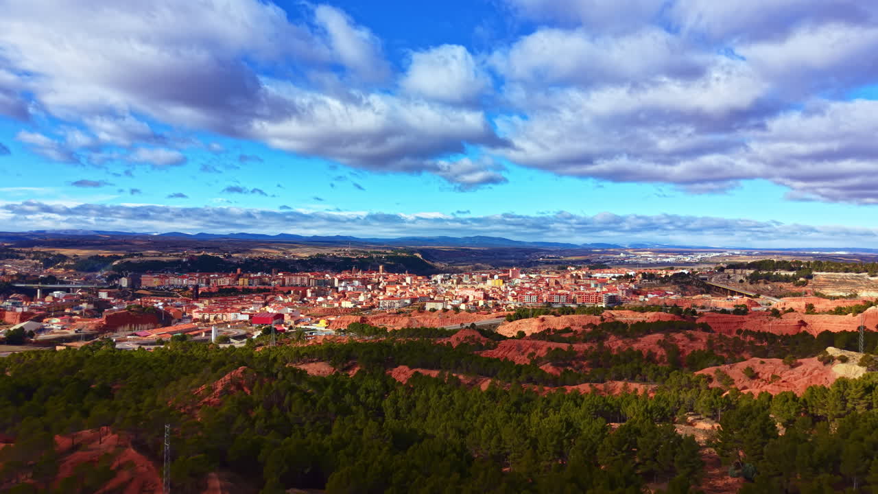 Vista di un paese con colline rosse e alberi verdi, sotto un cielo blu con nuvole sullo sfondo, di giorno, vicino alle montagne in Spagna.