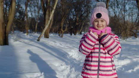 Una-Niña-Pequeña-Y-Alegre-Sonriendo,-Mostrando-Un-Gesto-De-Aprobación-En-Una-Carretera-Nevada-En-Un-Parque-De-Invierno-Al-Aire-Libre