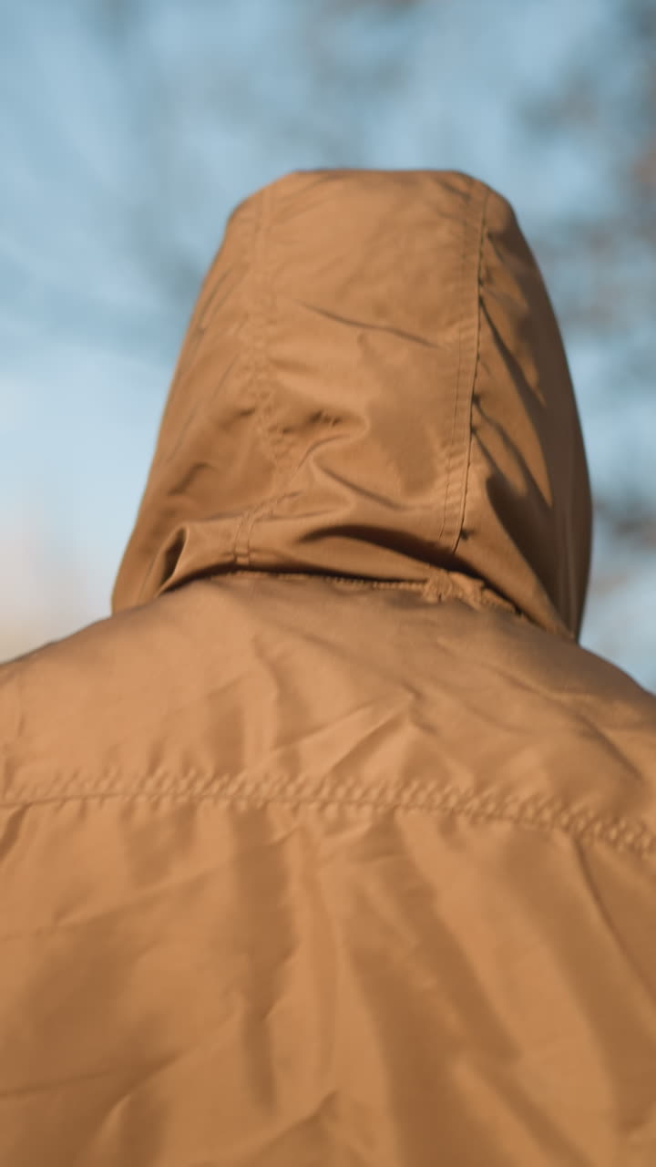 persona anónima caminando al aire libre en una chaqueta con capucha marrón contra un telón de fondo de árbol borroso, capturando una sensación de soledad y misterio