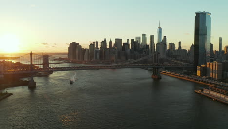 Forwards-fly-above-river.-Suspension-bridges-over-water-at-sunset.-Skyline-with-modern-tall-skyscrapers.-Heavy-traffic-on-roads.-Manhattan,-New-York-City,-USA