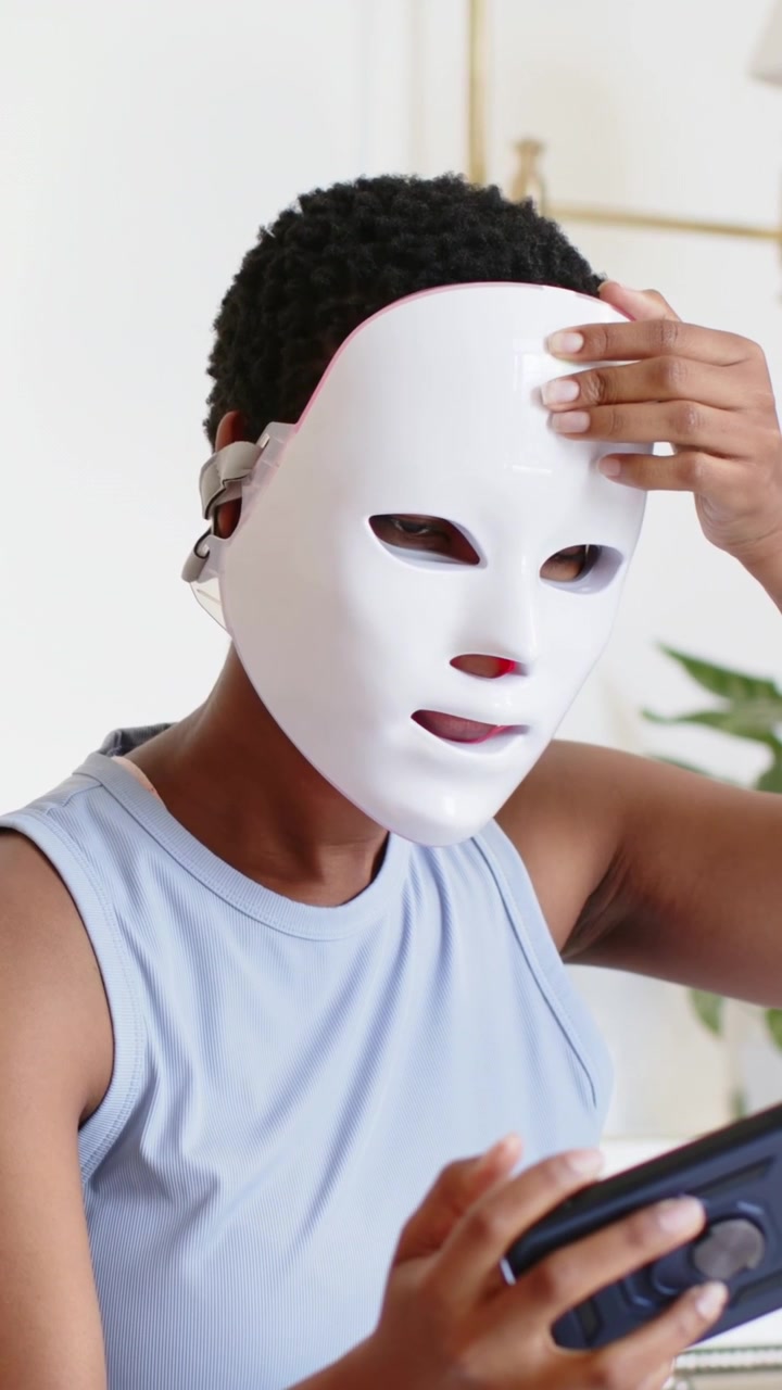 Vertikales Video: Frau in blauem Oberteil passt im Schlafzimmer eine LED-Maske an und hält eine Fernbedienung zum Starten bereit.