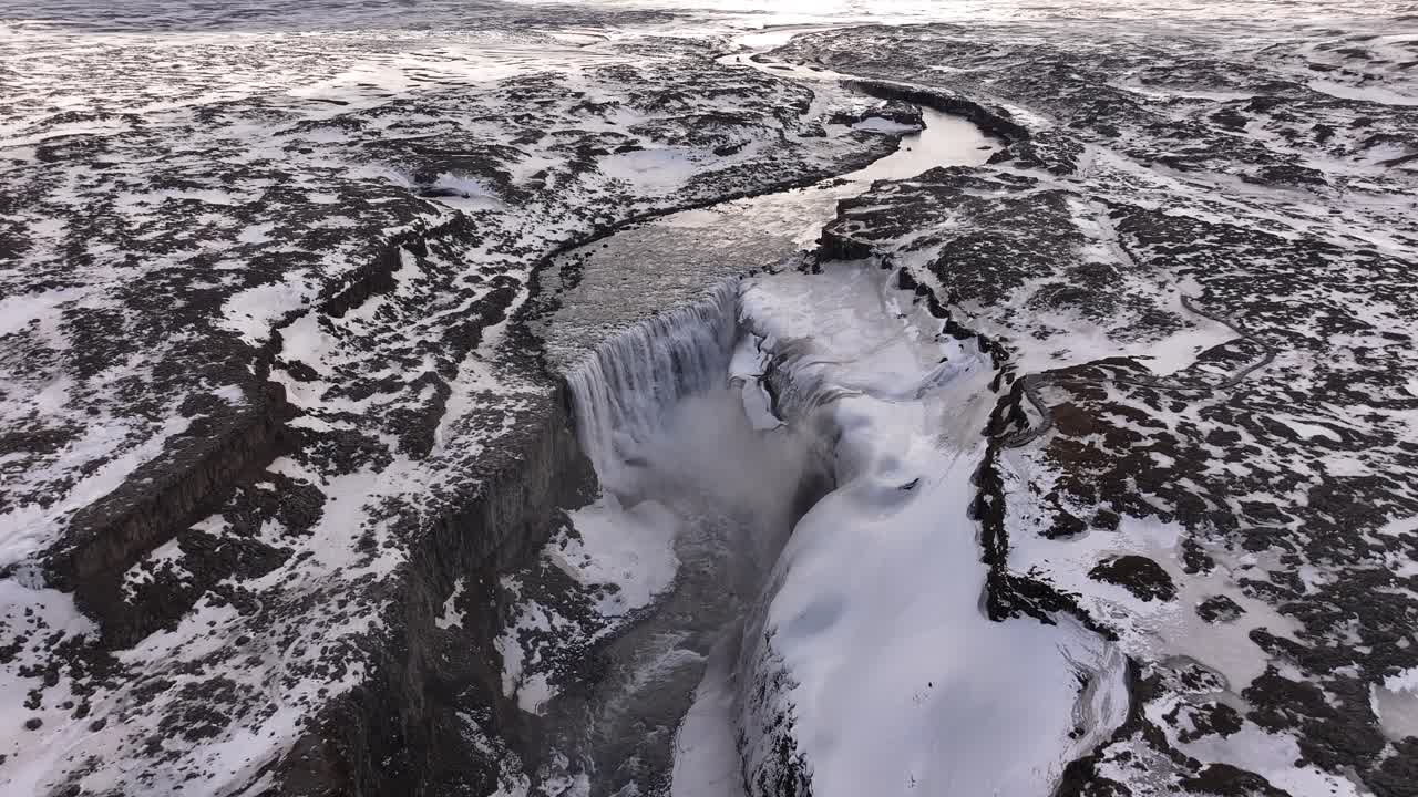 High-altitude aerial view of Dettifoss reveals the mighty waterfall carving through Iceland's frozen volcanic plateau, with the river winding across a stark snow-covered landscape.
