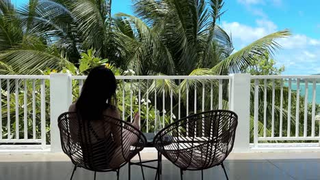 back view of girl in black swimsuit sitting on luxurious balcony admiring palm tree view on sunny day