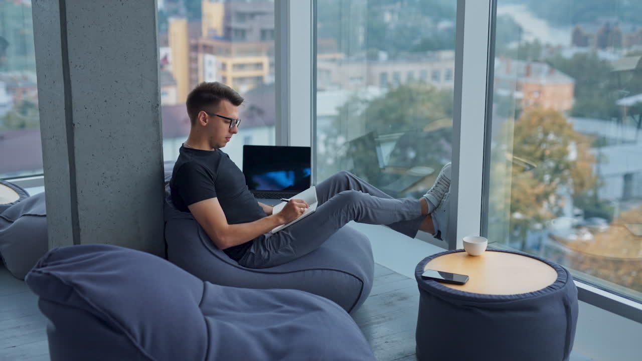 Coming closer to a young man sitting relaxed near the window. Man wearing glasses writing something in paper book. Blurred backdrop.